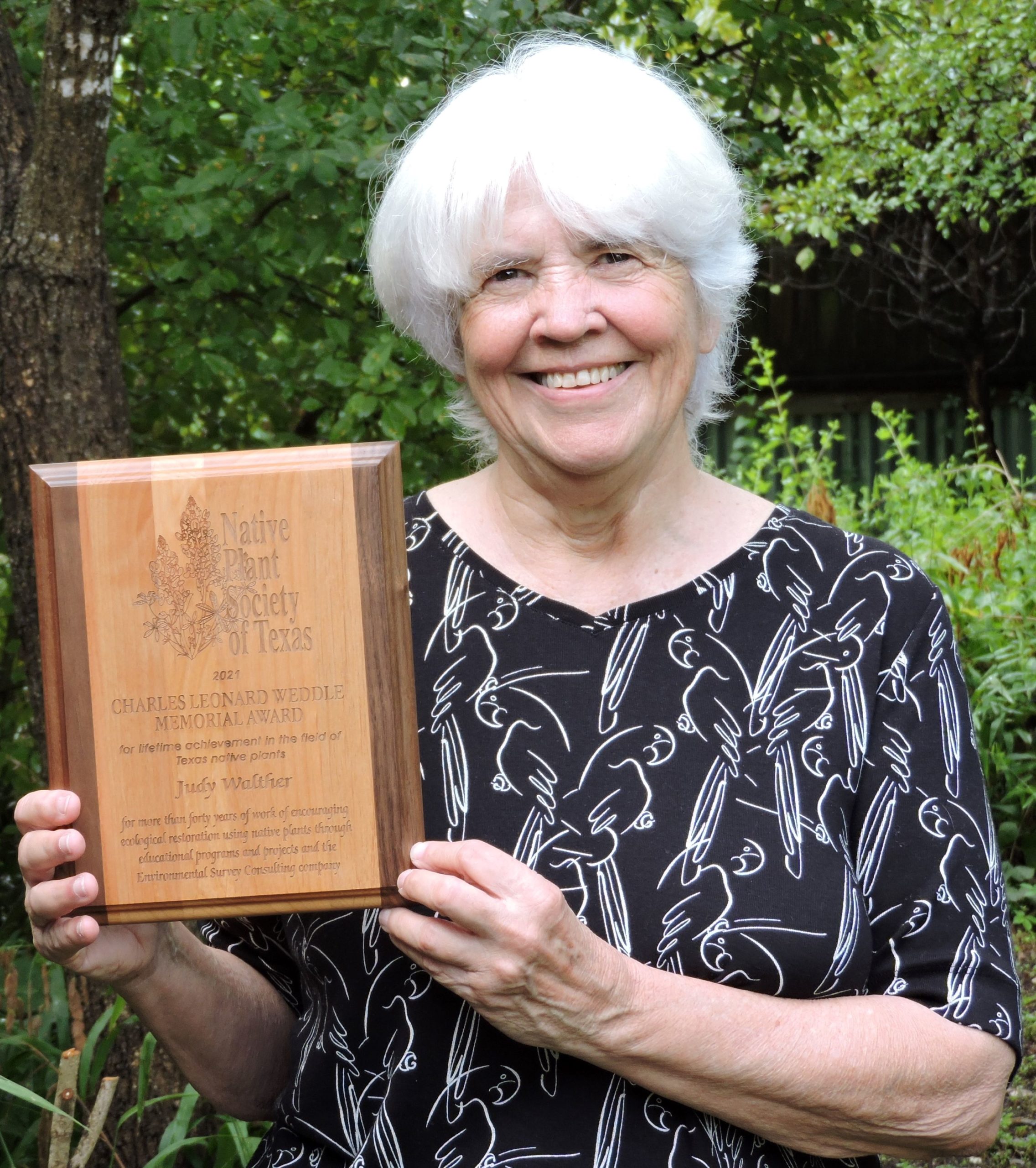 Judy Walther standing with NPSOT award in front of native trees and plants